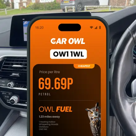 A mobile phone displaying the Car Owl app, showing petrol price per litre as 69.69p and the location OW1 1WL, with a steering wheel and car dashboard in the background.