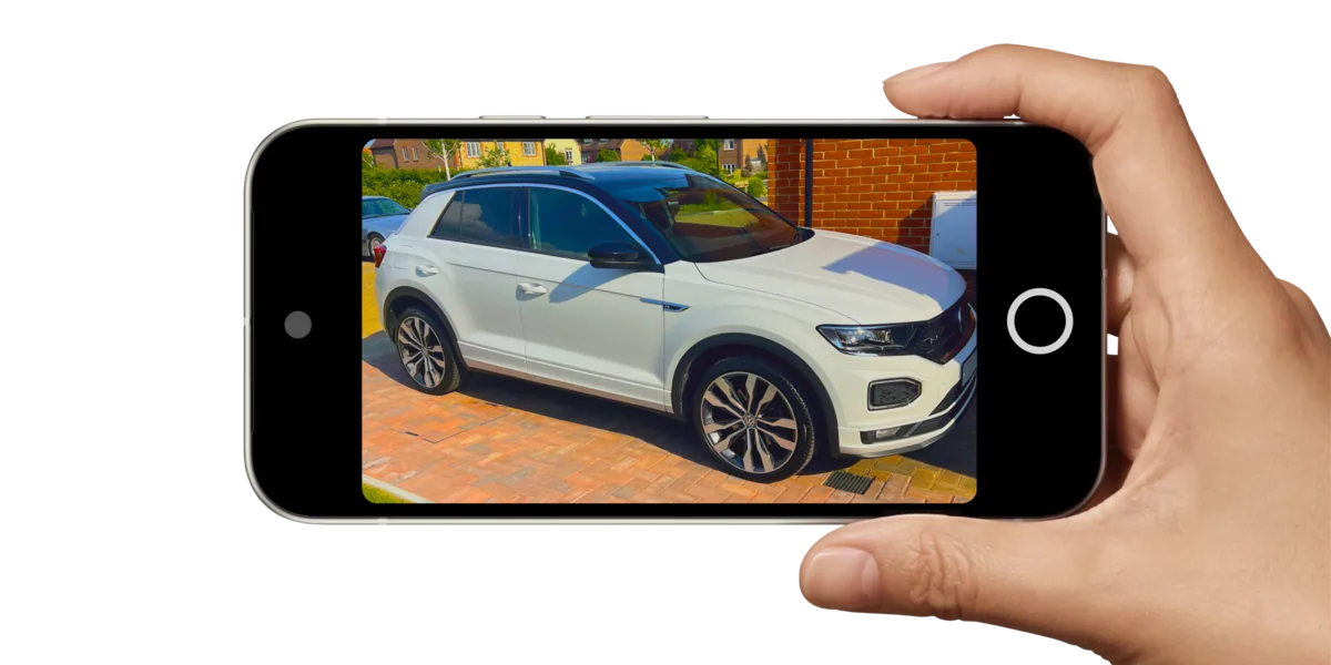 A hand holding a smartphone displaying a photo of a white SUV parked on a brick driveway in front of a house.